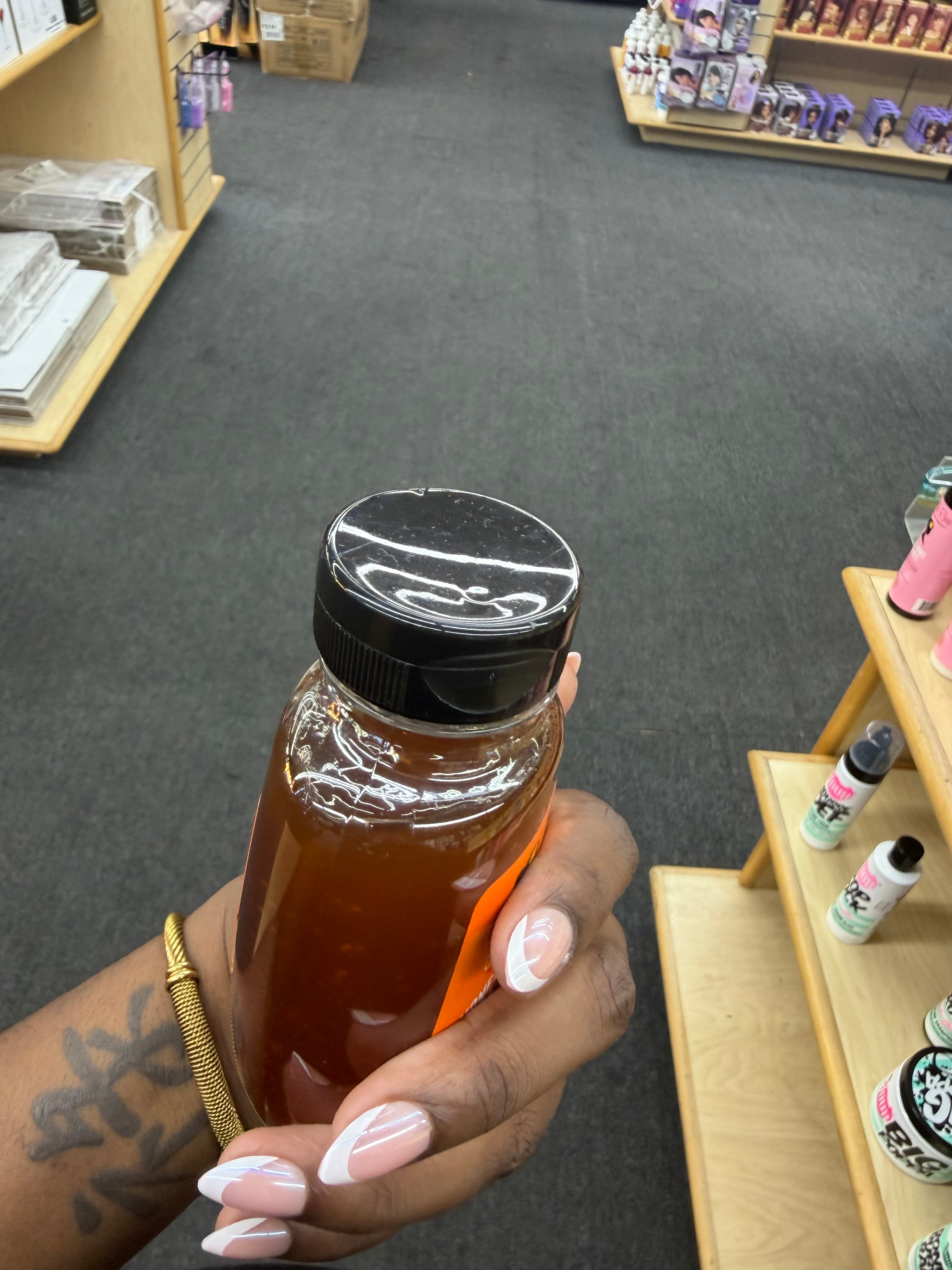 A hand with pink and white French-tip nails holds a clear plastic squeeze bottle filled with amber honey in a retail store setting.