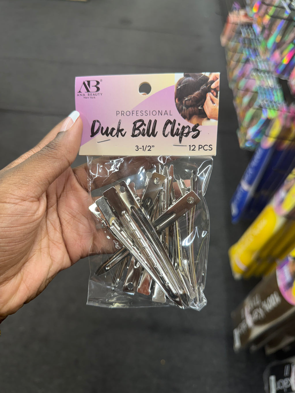 A hand holding a clear plastic bag of AB All Beauty Professional Duck Bill Clips, 12 pieces, size 3-1/2 inches, against a blurred store background.