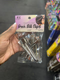 A hand holding a clear plastic bag of AB All Beauty Professional Duck Bill Clips, 12 pieces, size 3-1/2 inches, against a blurred store background.