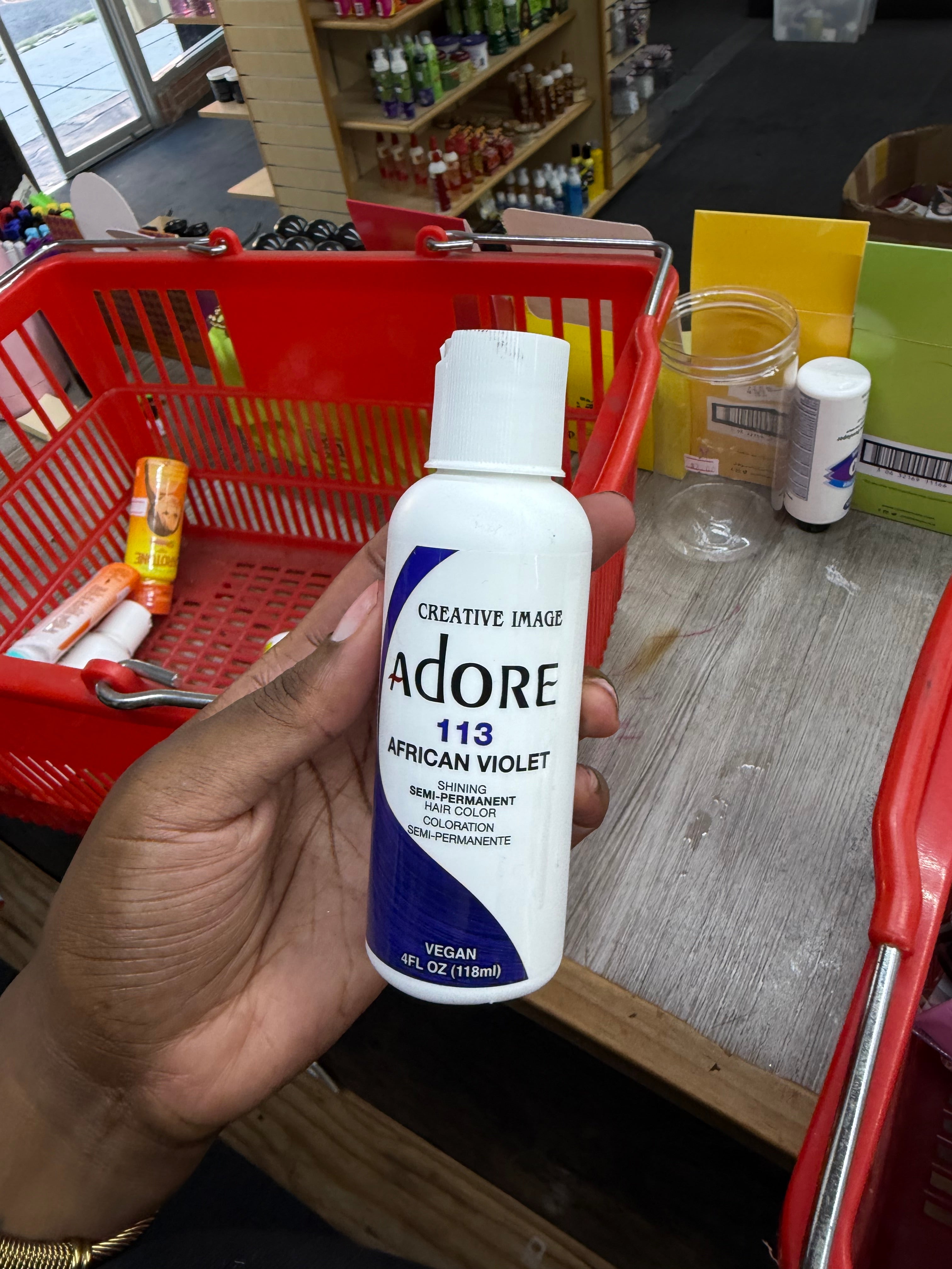 A hand holds a bottle of Creative Image Adore 113 African Violet photography and makeup medium in a grocery store checkout area, with a red shopping basket and store shelves visible in the background.