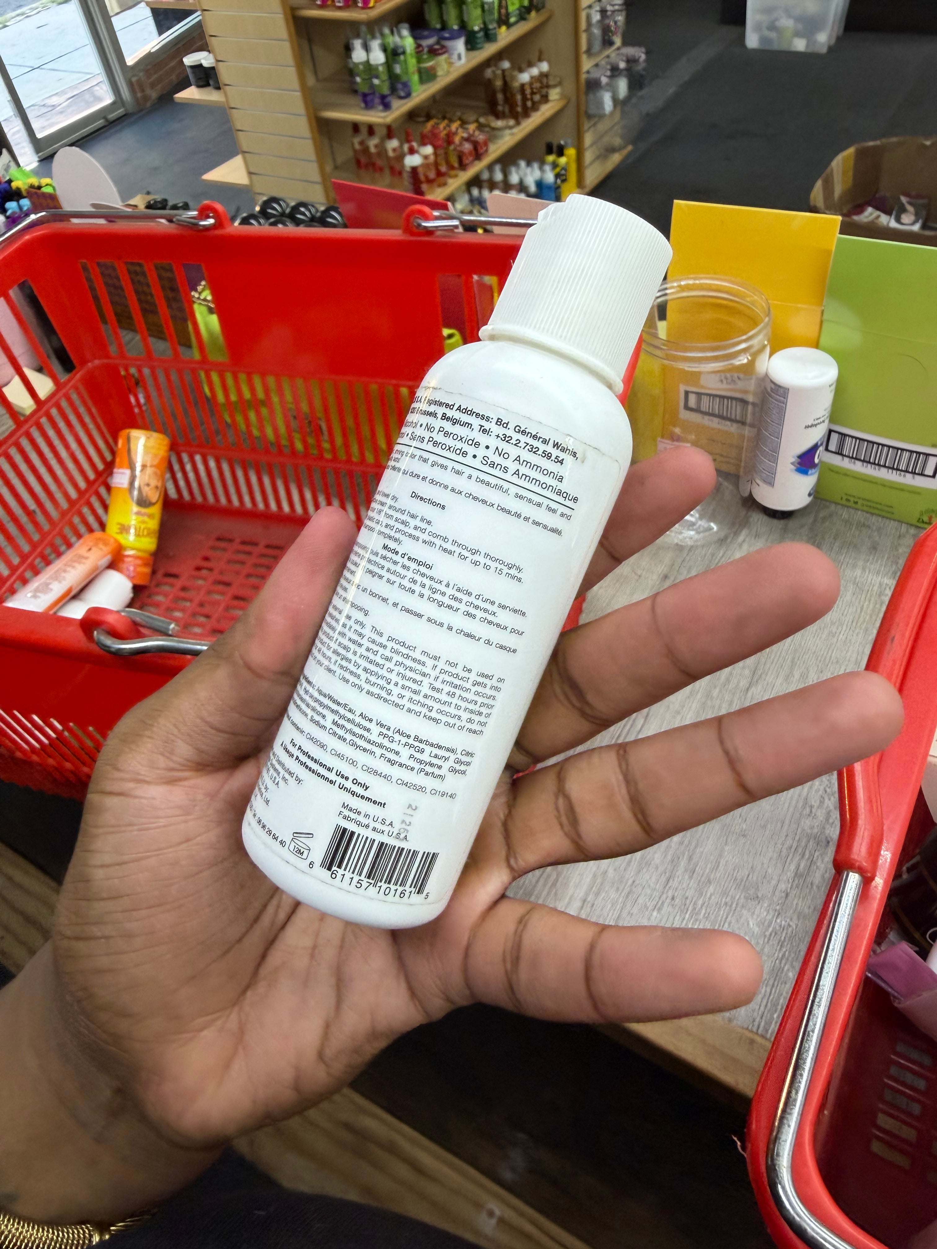 A hand holding a white plastic bottle with a white cap, showing the back label, in a store setting with a red shopping basket and other products in the background.