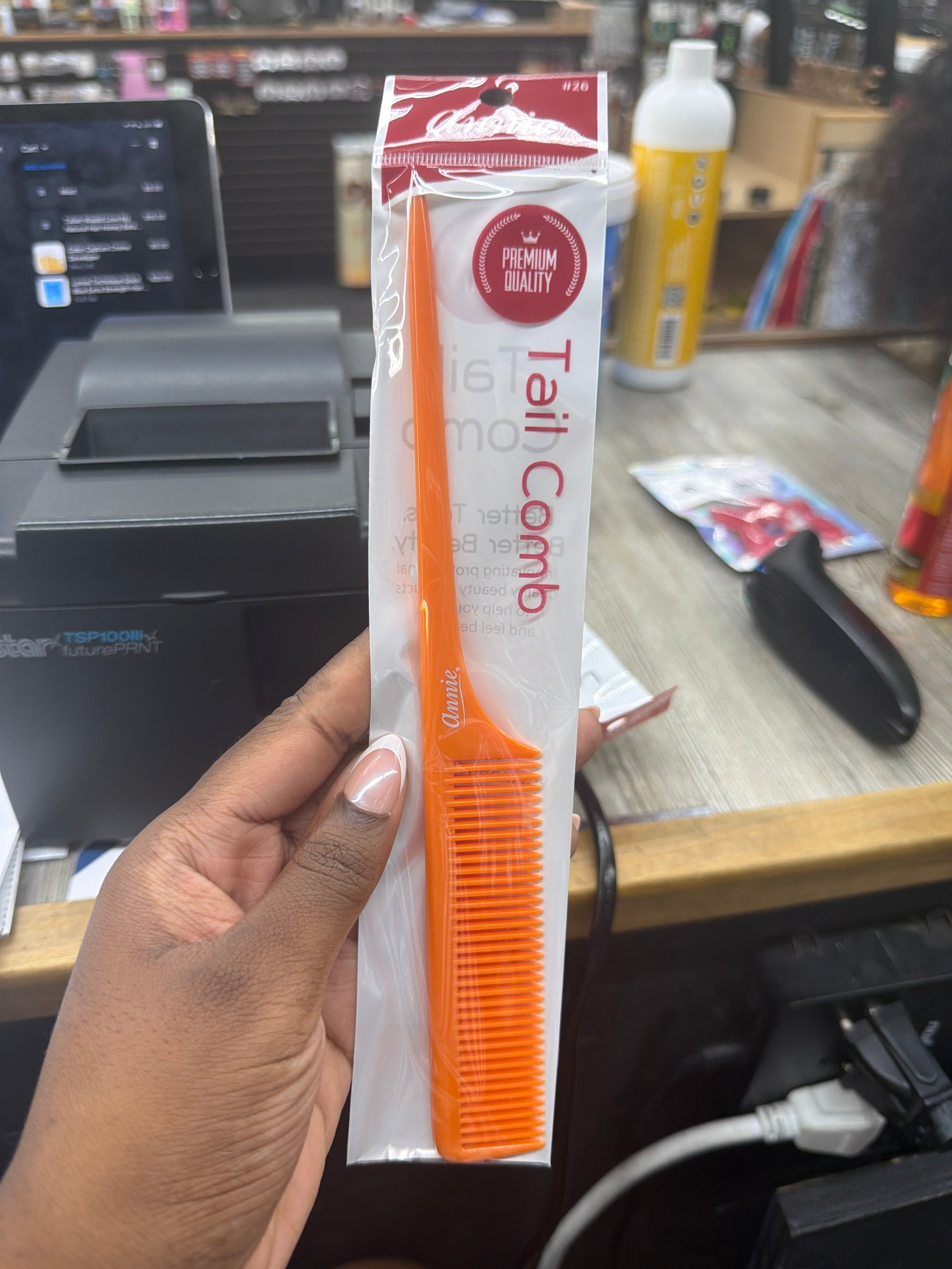An orange plastic tail comb in its white and red retail packaging, held by a hand in front of a busy retail counter.