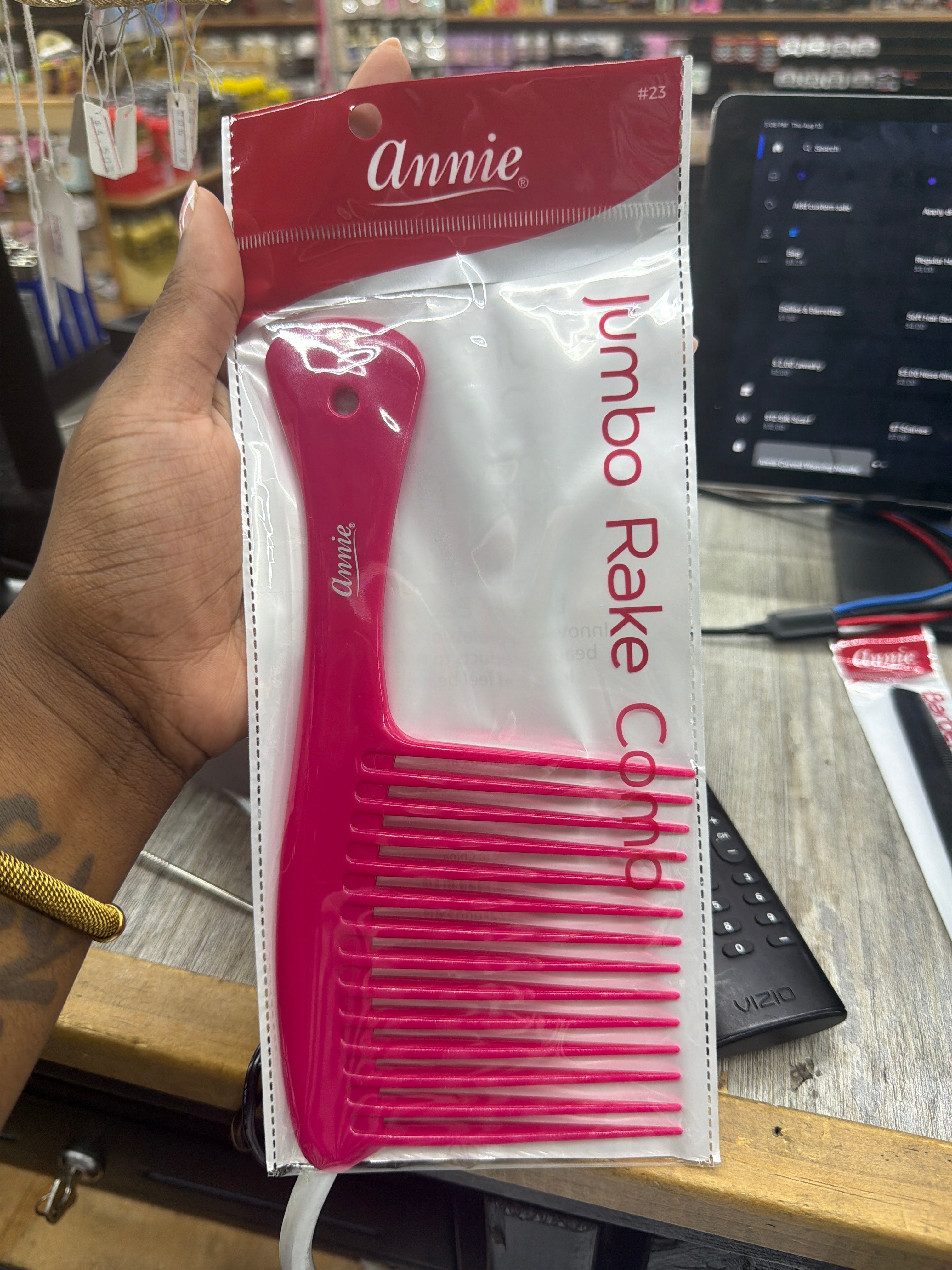 A hand holds a packaged bright pink Annie Jumbo Rake Comb in a clear plastic bag with a red header, photographed in a retail or office setting with a laptop and desk visible in the background.