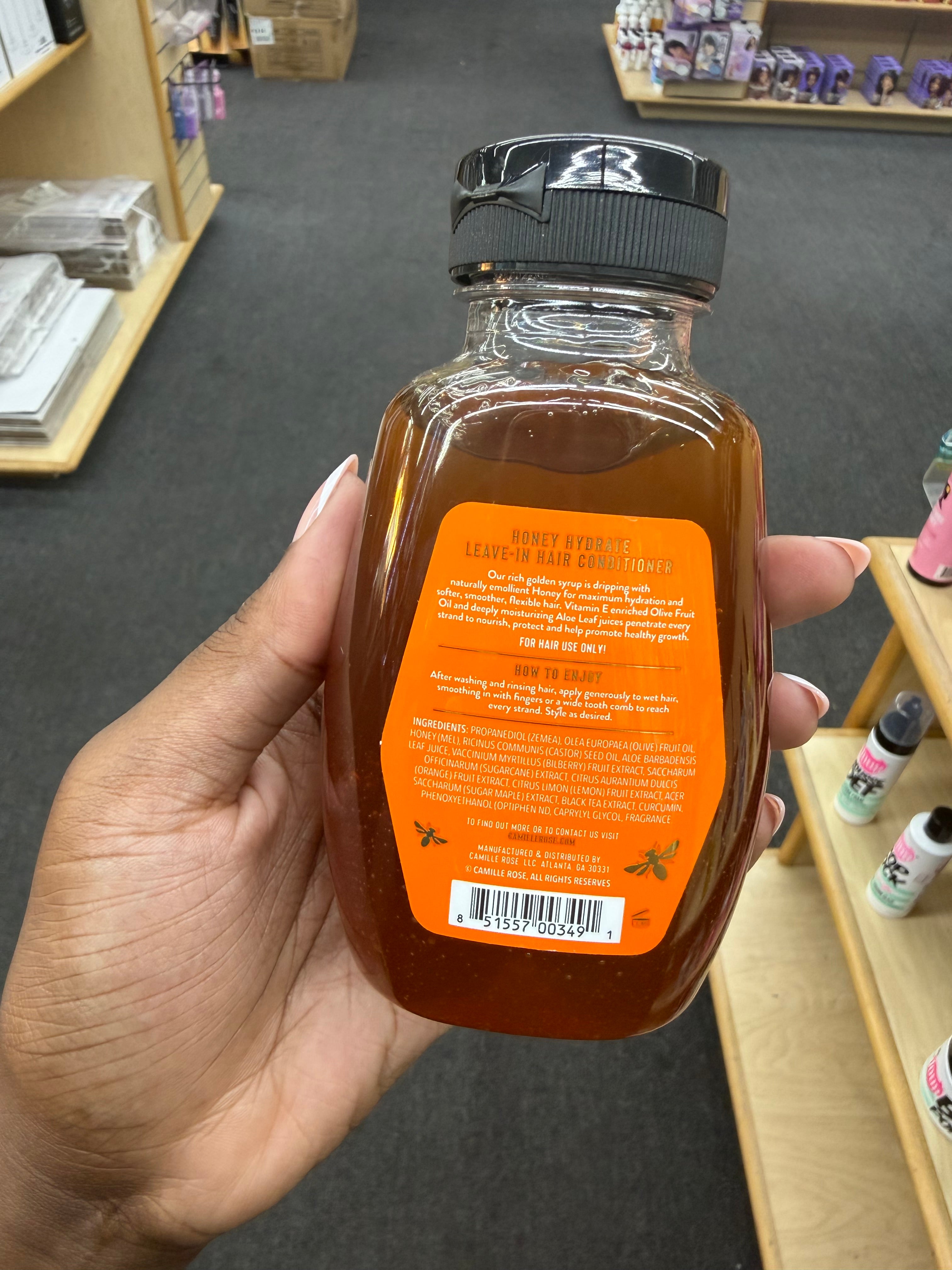 A hand holds a bottle of honey in a retail store. The bottle is clear glass filled with amber-brown honey and has a black cap.