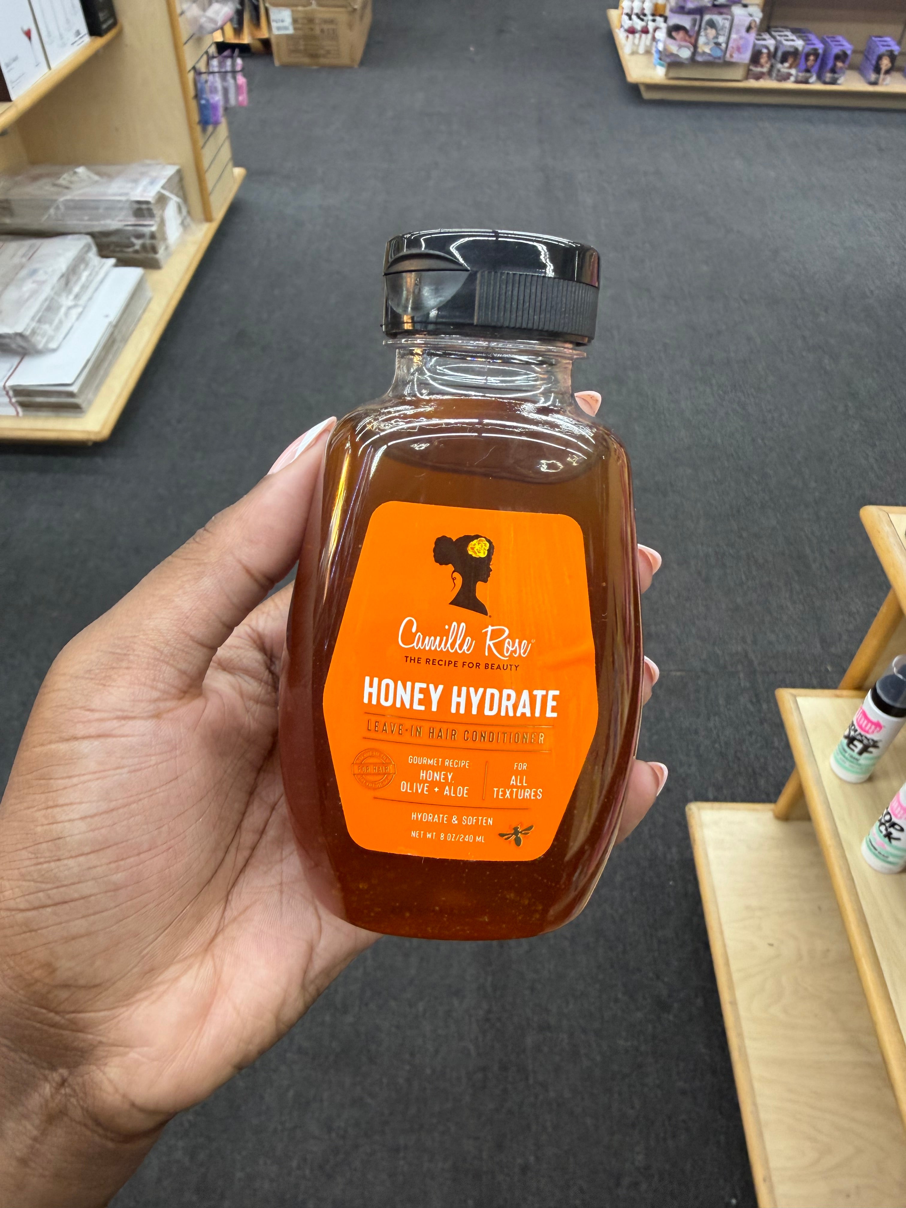 A hand holds a bottle of Candit Rose Honey Hydrate in a retail store setting. The bottle is made of clear glass, showing the amber-brown honey inside, and has a prominent orange label with a black cap.