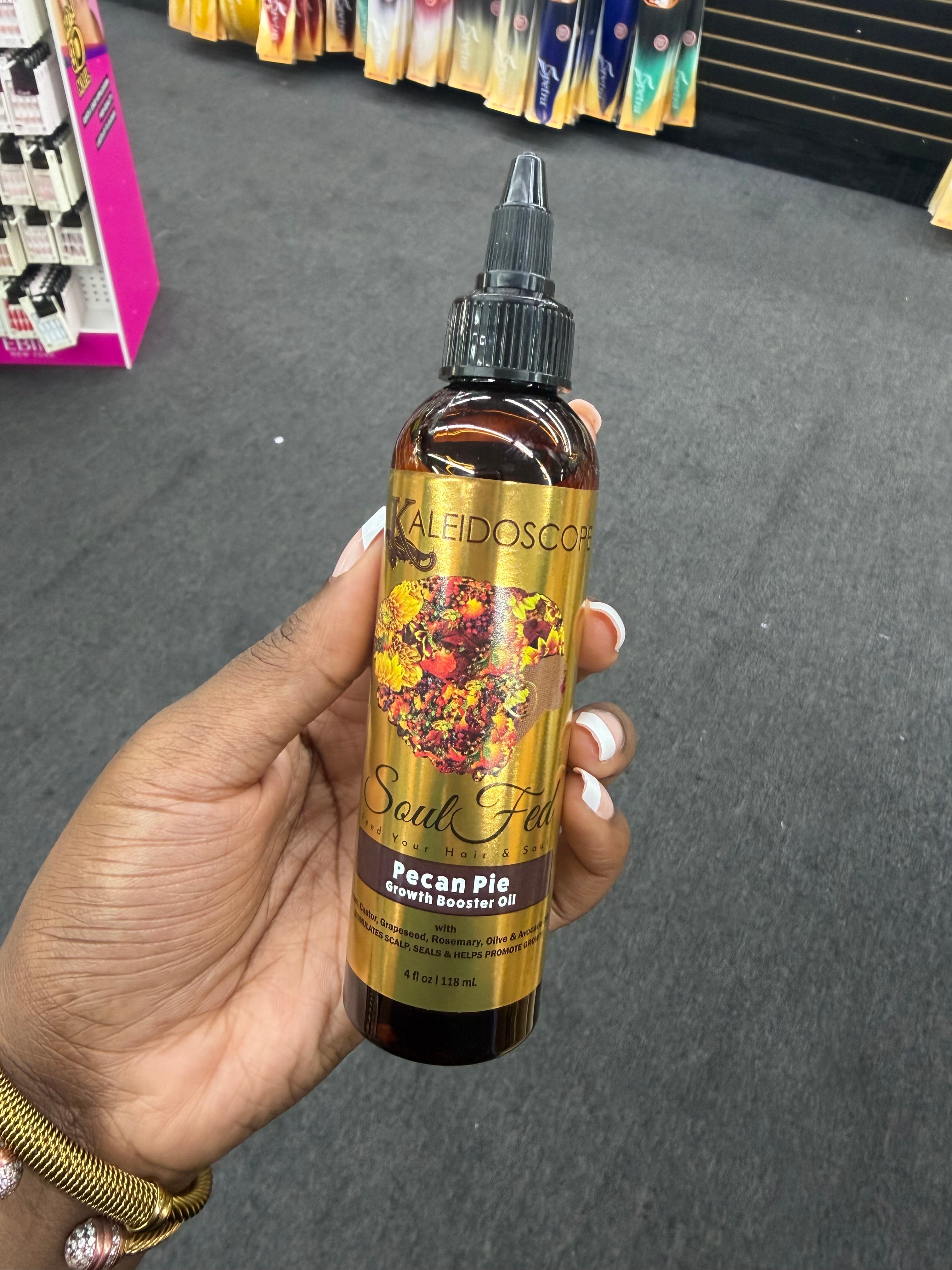 A hand holds a bottle of Kaleidoscope Soul Fx Pecan Pie Growth Booster Oil against a dark gray carpeted background.