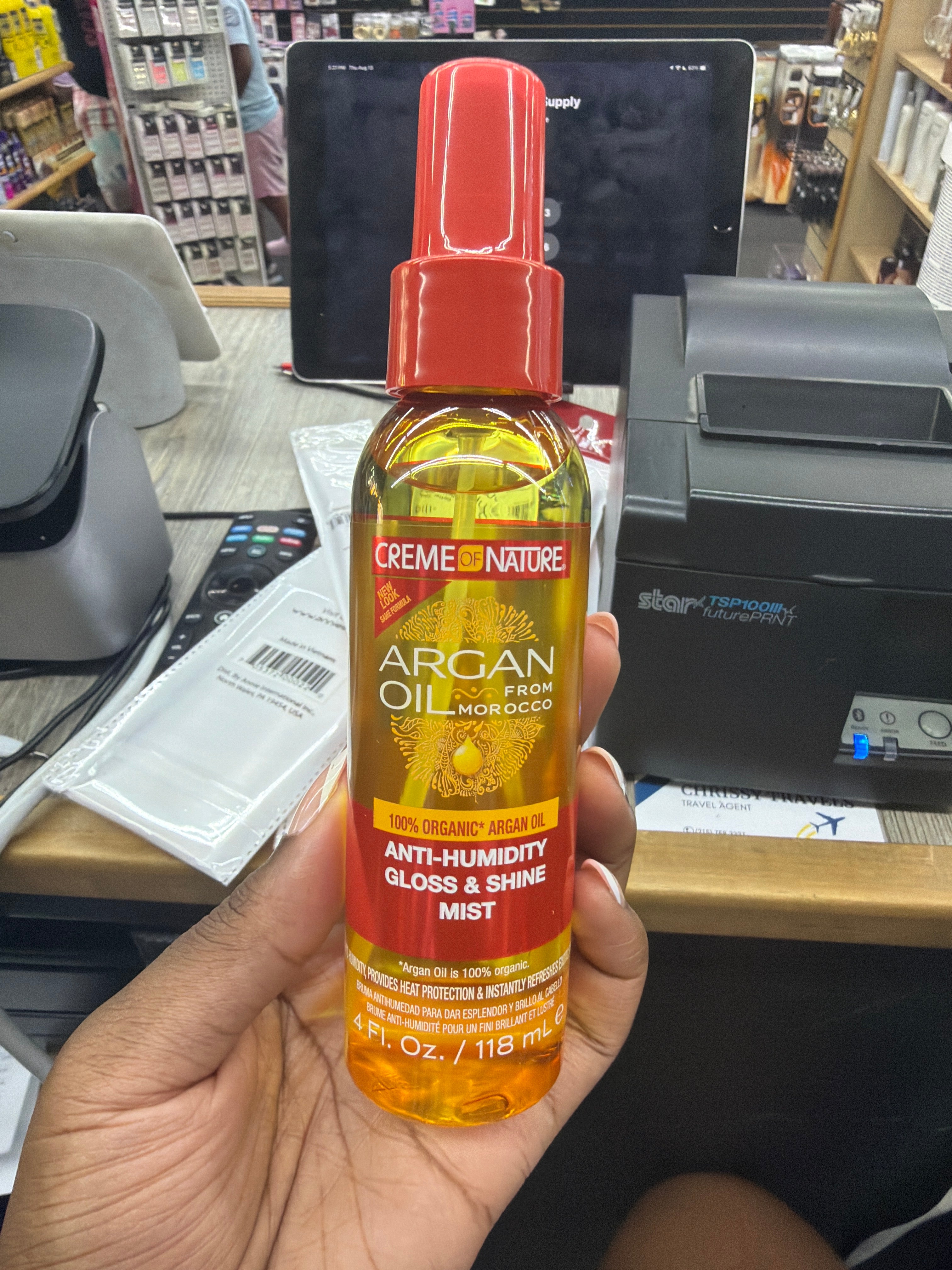 A hand holding a bottle of Creme Nature Argan Oil Anti-Humidity Gloss & Shine Mist in a retail store setting.