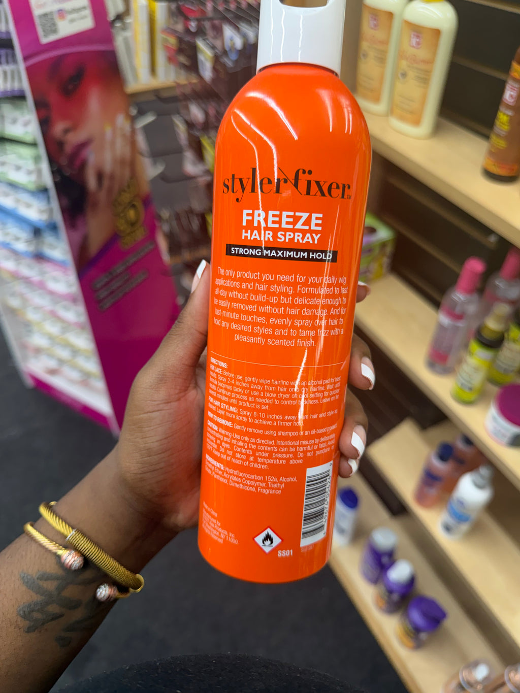 A hand holding an orange bottle of StylerFixer Freeze Hair Spray in a retail store setting with shelves of other hair products in the background.