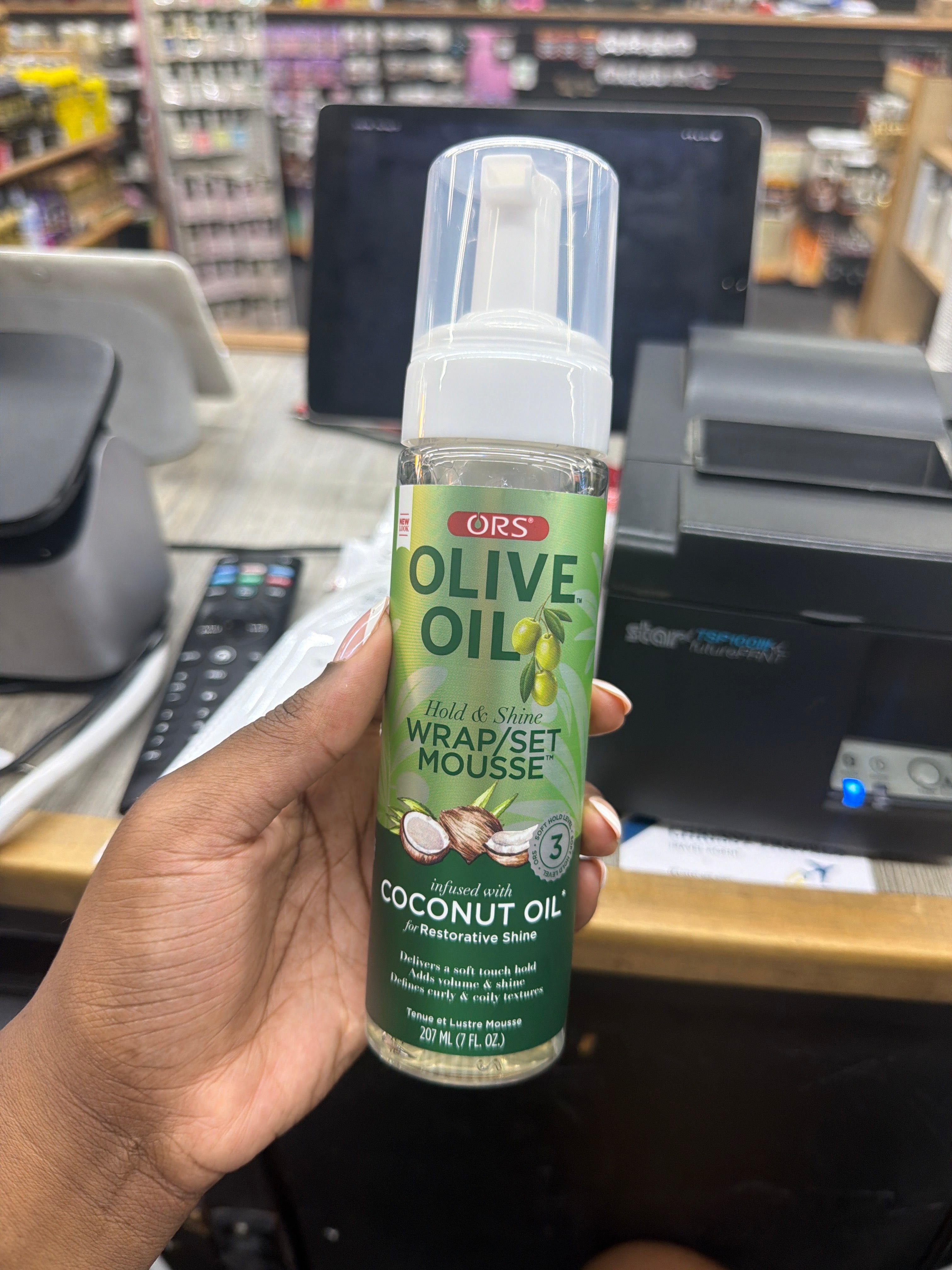 A hand holding an ORS Olive Oil Hold & Set Wrap/Set Mousse bottle in a retail store setting with a cash register and shelves in the background.
