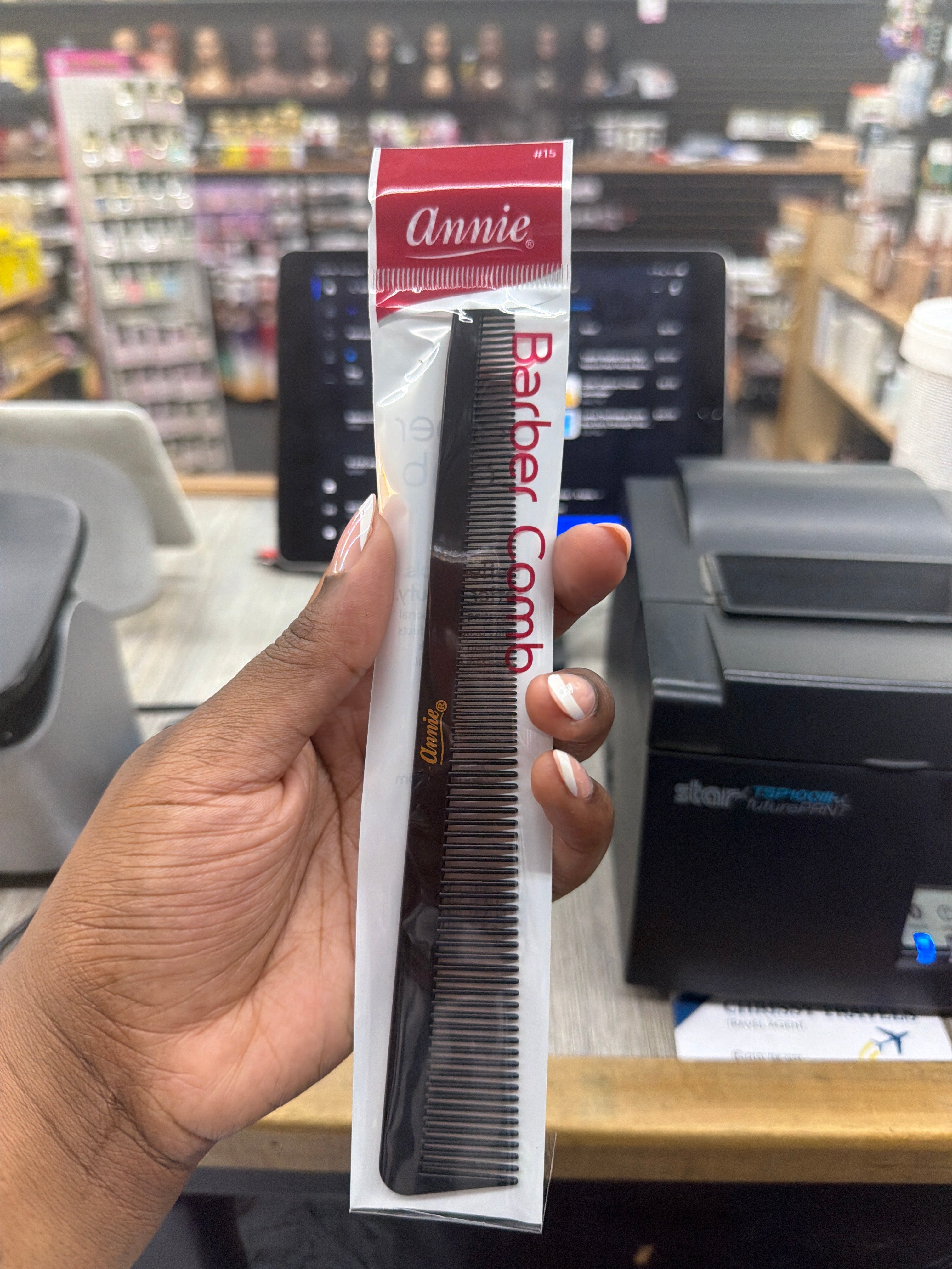 A hand holds a packaged black barber comb in a retail store setting. The packaging has a red top label that reads 'Annie' and the comb itself is black with gold lettering.