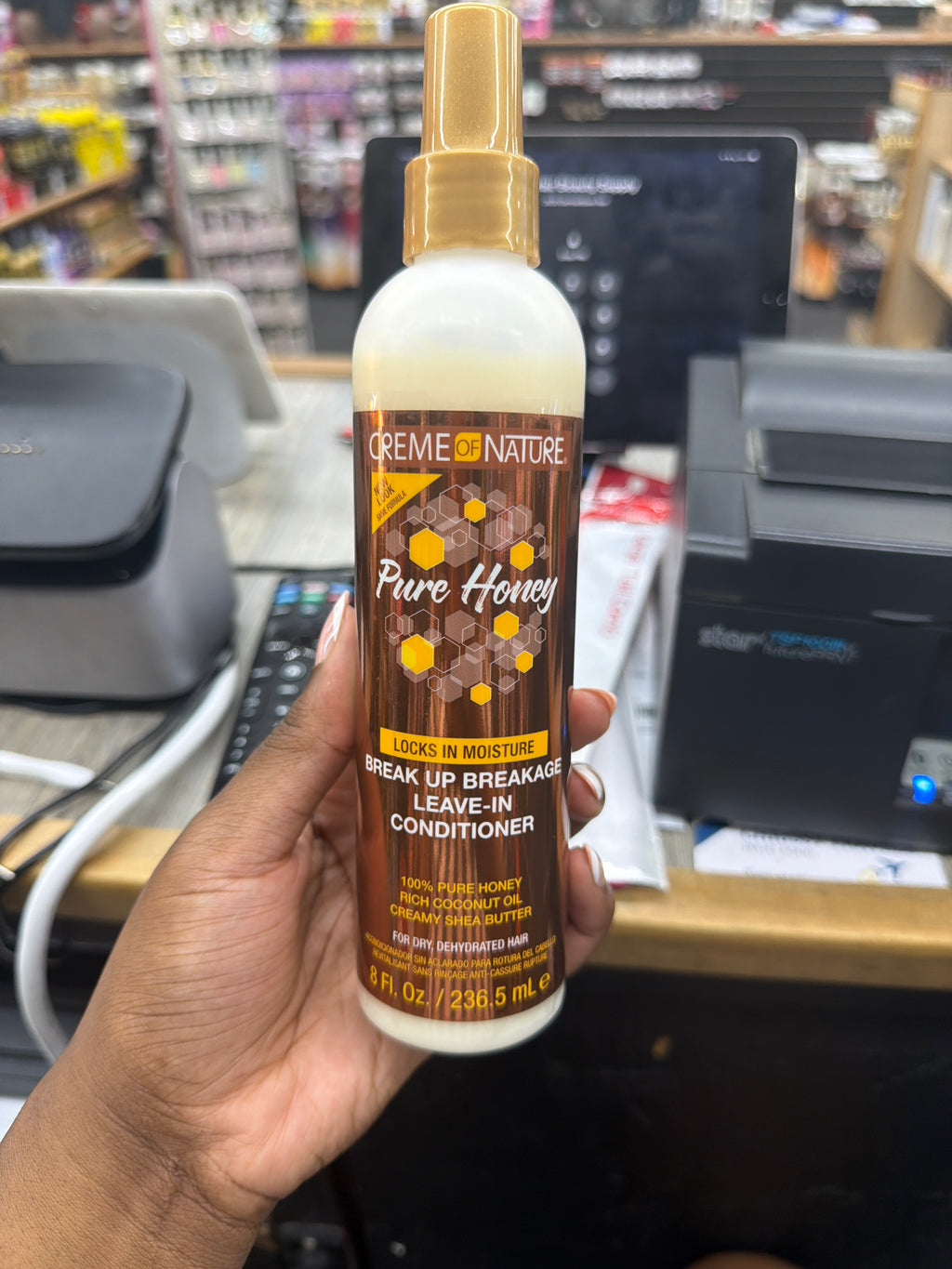 A hand holding a bottle of Creme de Nature Pure Honey Leave-In Conditioner in a retail store setting with a cash register in the background.
