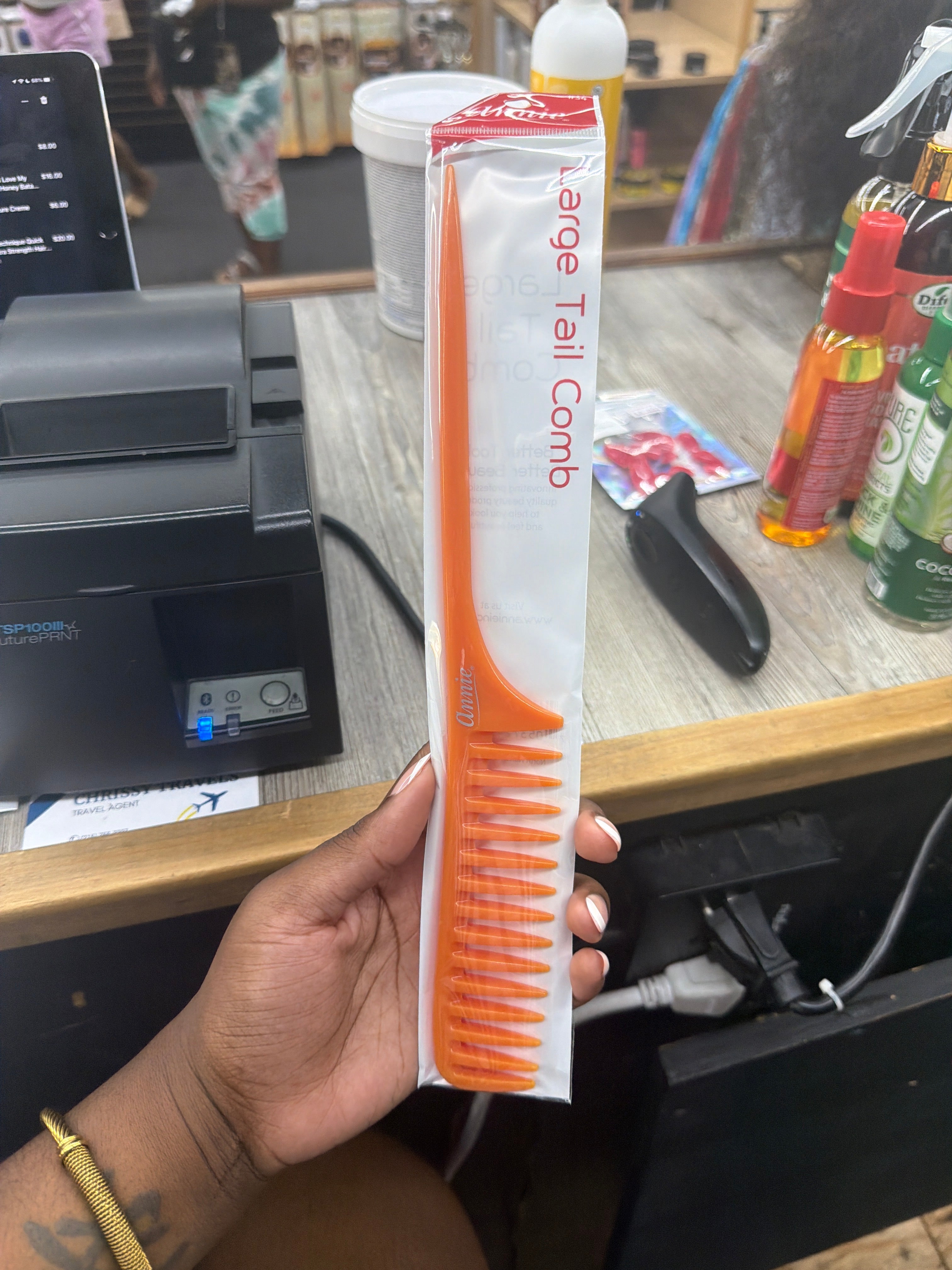 An orange hair tail comb in its white and red retail packaging, held by a hand in a retail store setting.