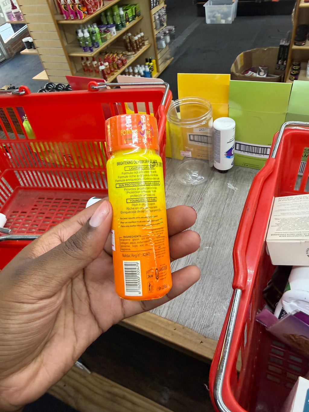 A hand holding a small orange and yellow bottle with its label facing away from the camera, set in a busy retail store environment with red shopping baskets and shelves in the background.