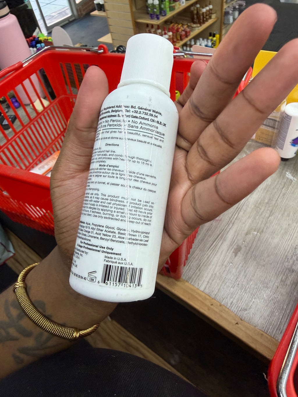 A hand holding a white plastic bottle with a white cap, showing the back label, in a store setting with a red shopping basket and wooden counter.