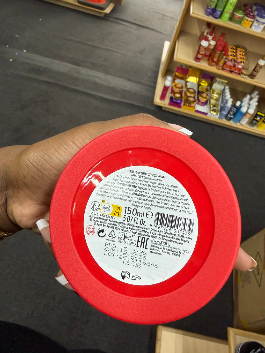 A hand holding the bottom of a red plastic jar, showing the white product information label with a barcode and text. The background shows a store shelf with other cosmetic products.