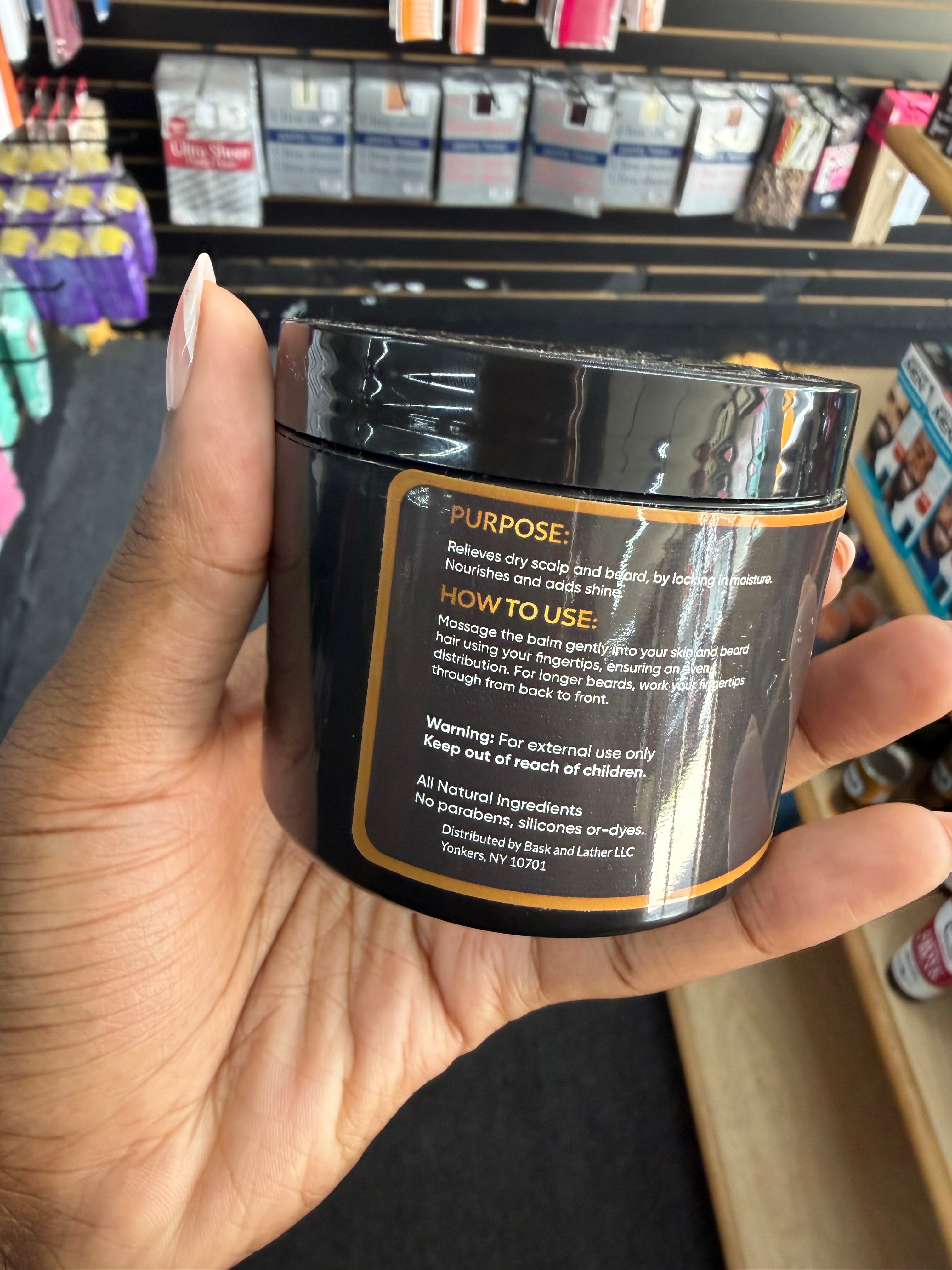 A hand holding a black jar of hair product in a retail store setting, showing the back label with text including 'PURPOSE' and 'HOW TO USE'.