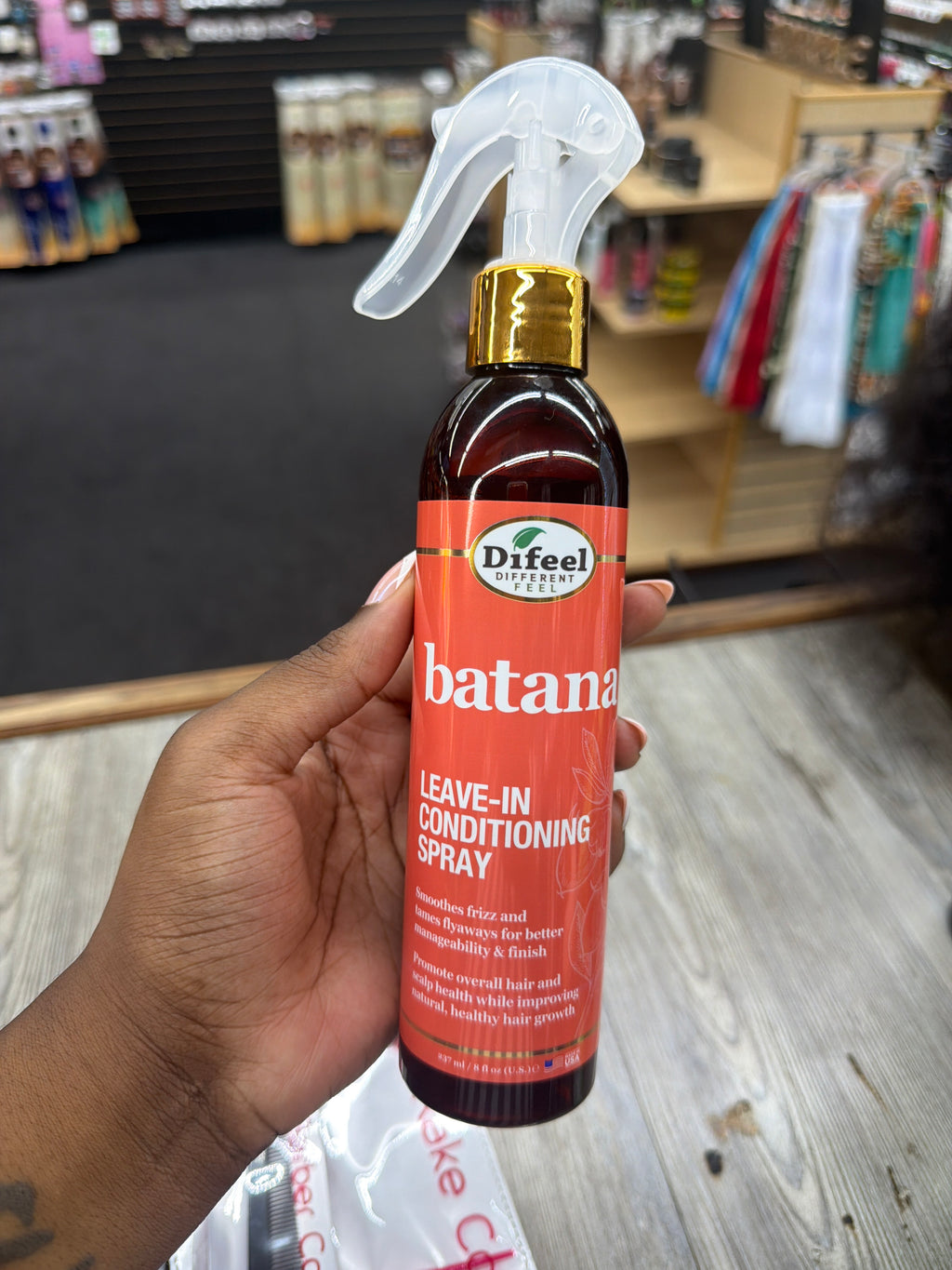 A hand holding a bottle of Difeel Naturals Batana Leave-In Conditioning Spray in a retail store setting.