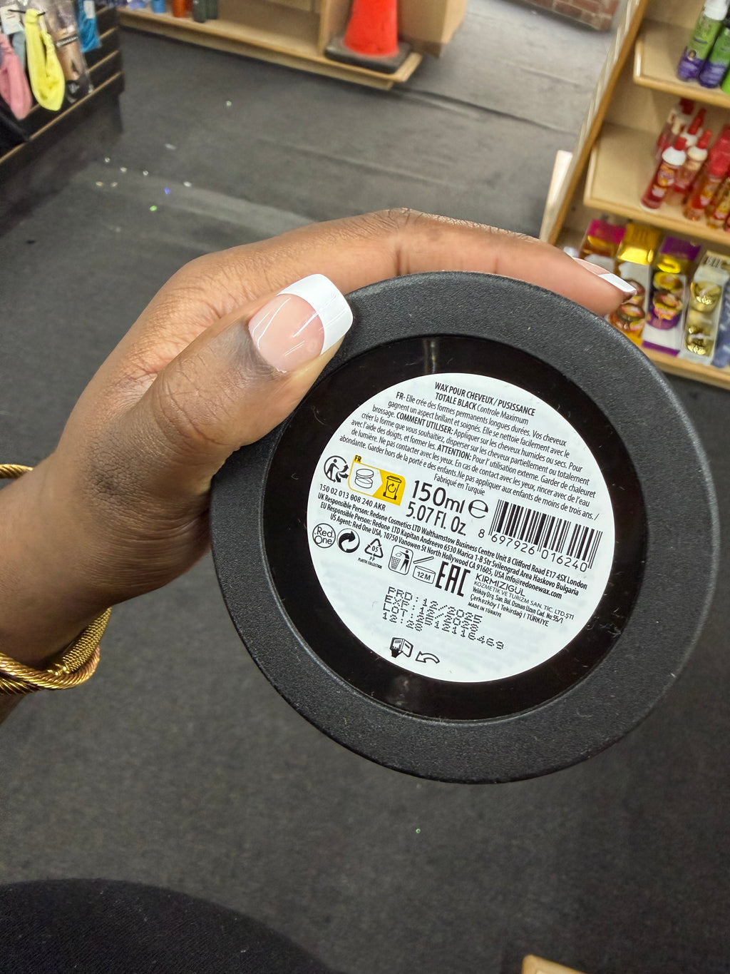 A hand holding the bottom of a round black cosmetic jar, showing the white product label with text and barcode. The background shows a store interior with shelves and products.