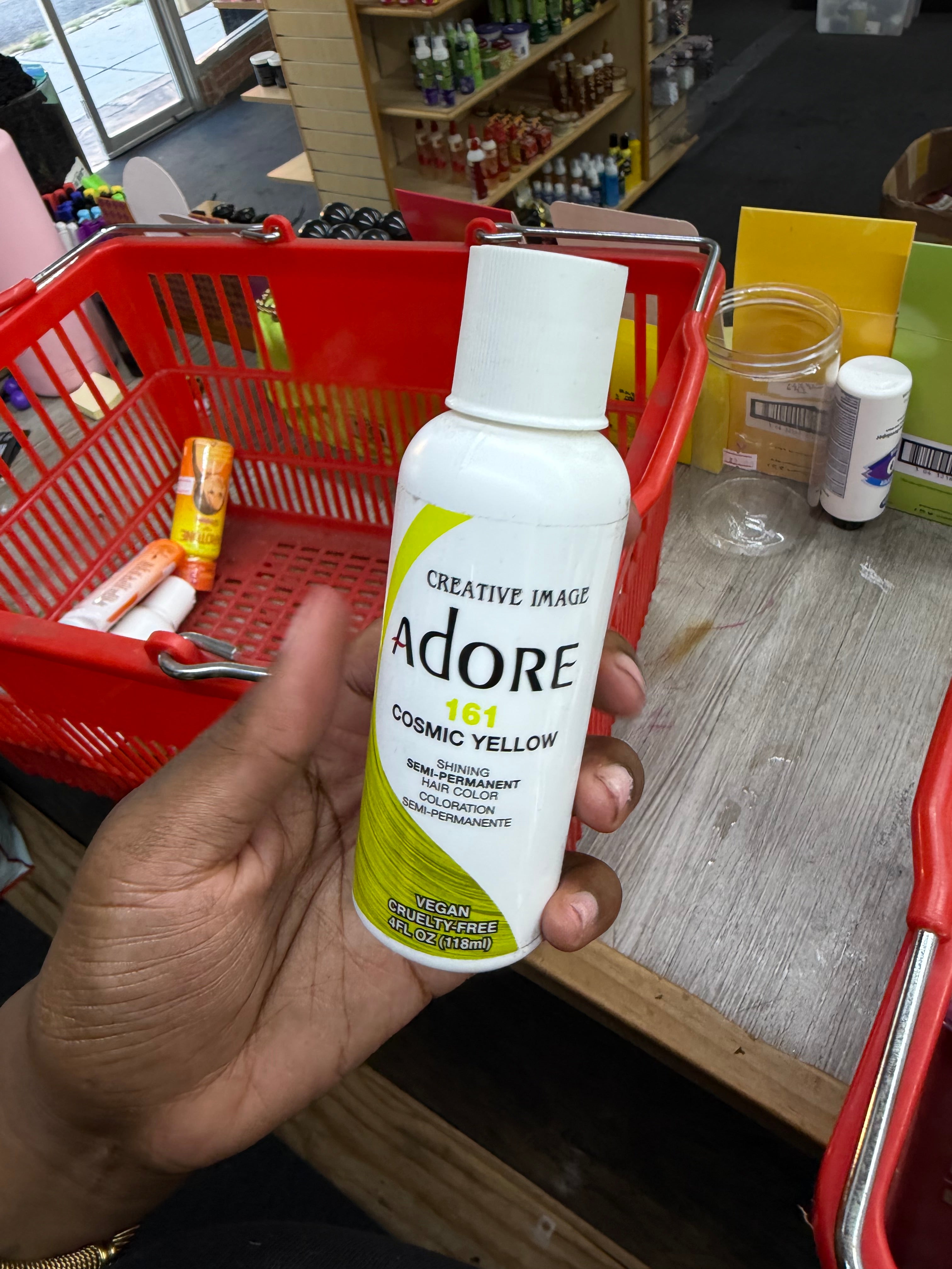 A hand holding a white bottle of Creative Image Adore 161 Cosmic Yellow cosmetic pigment in a retail store setting with a red shopping basket in the background.