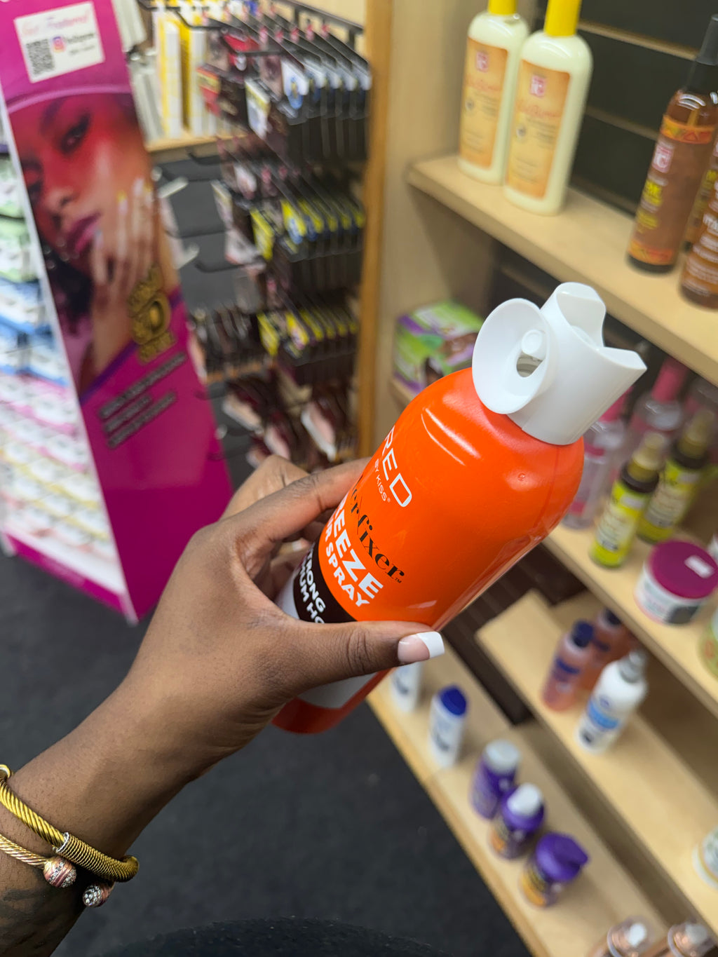 A hand holding an orange bottle of hair product with a white flip-top cap, photographed in a retail store aisle with shelves of other products in the background.