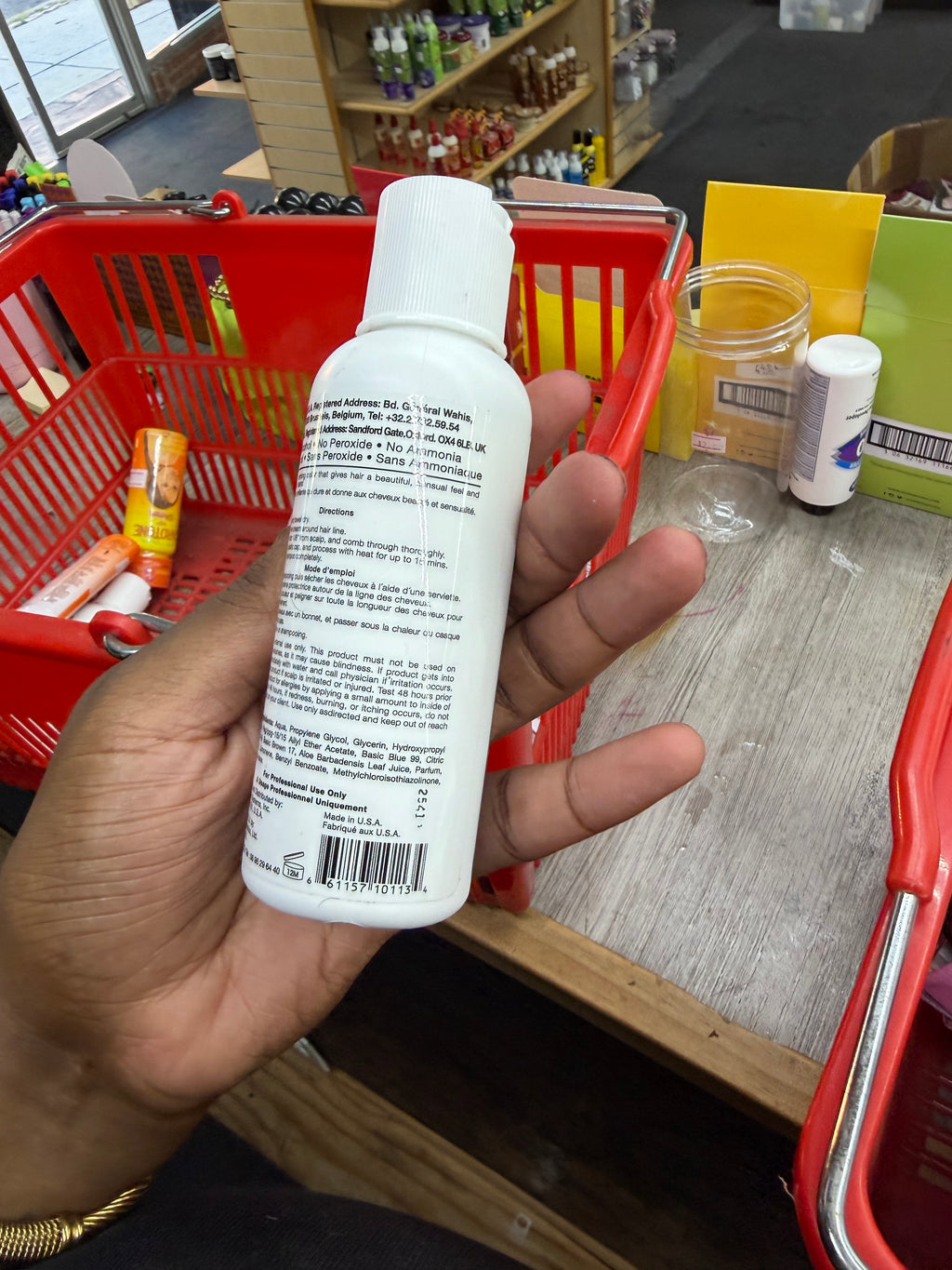 A hand holding a white plastic bottle with a white cap, showing the back label, in a store setting with red shopping baskets and a counter.