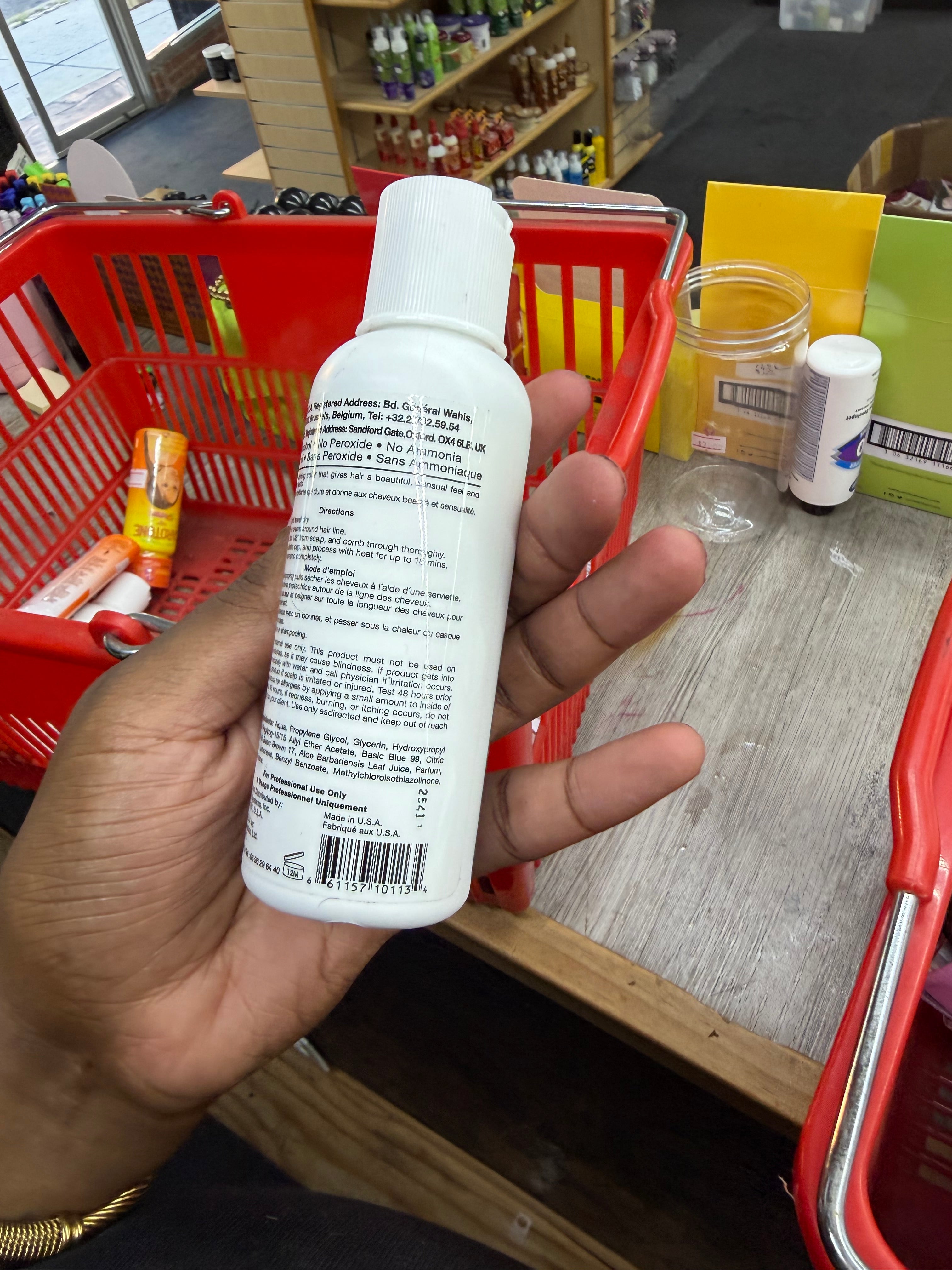 A hand holding a white plastic bottle with a white cap, showing the back label, in a store setting with red shopping baskets and a counter.