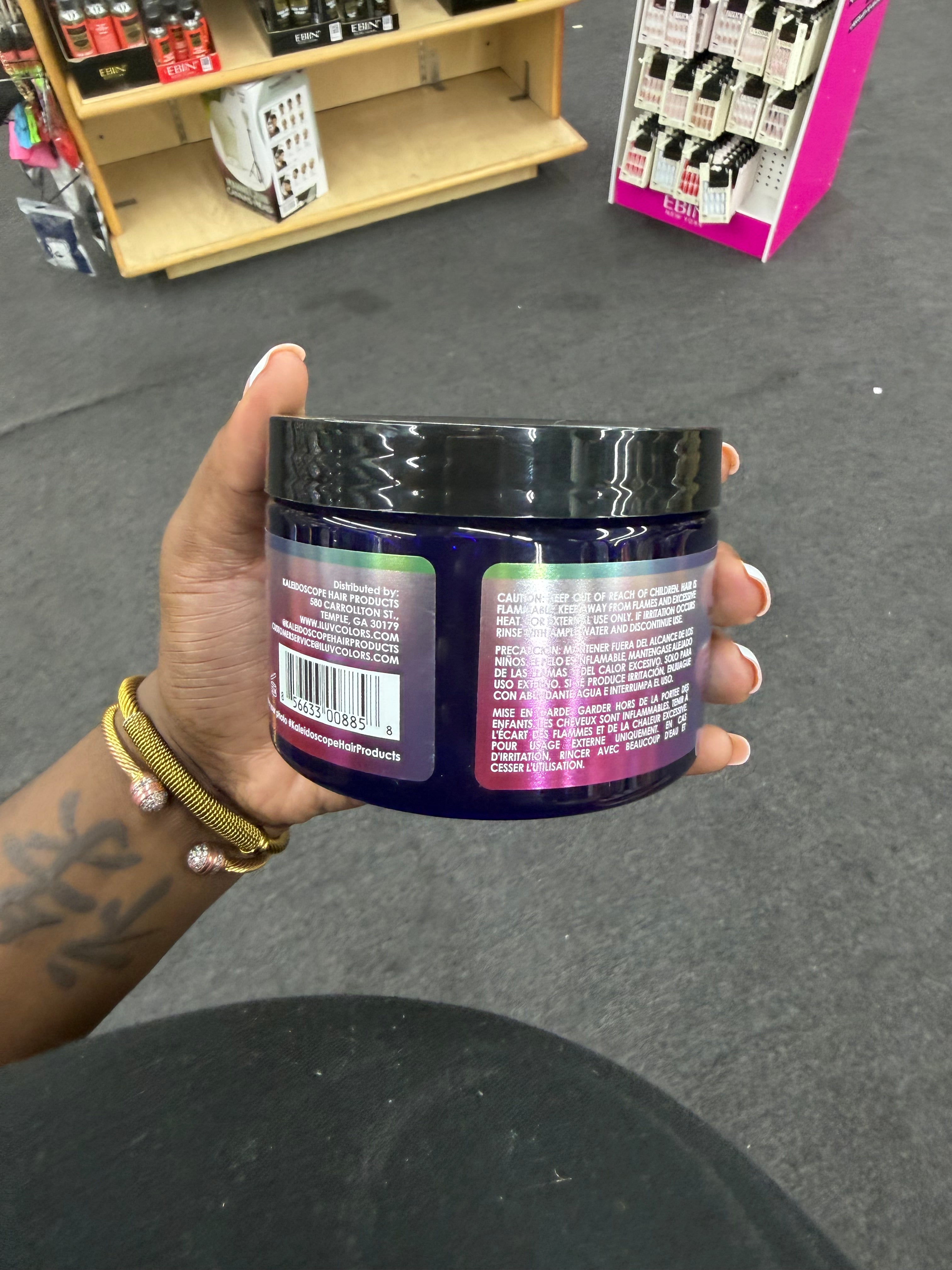 A hand holding a dark purple jar of hair product with a black lid, showing the back labels, in a store setting with shelves and a gray floor in the background.