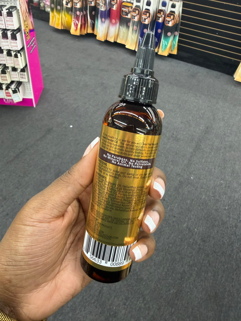 A hand holding a brown amber dropper bottle with a black cap and a gold label, photographed in a retail store setting with a gray carpet and product displays in the background.