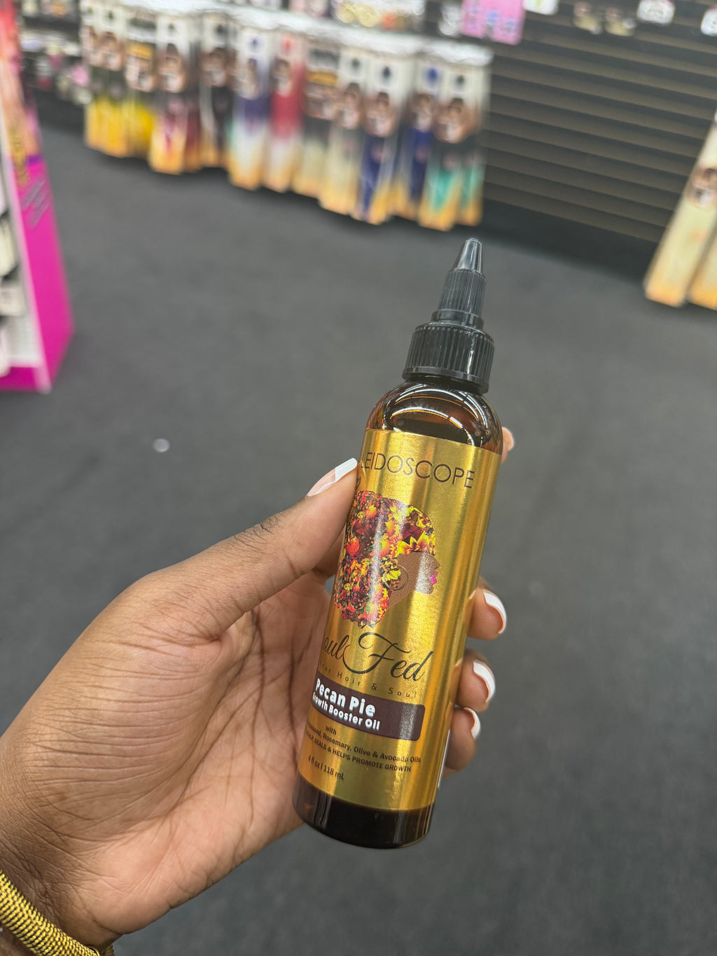 A hand holding a bottle of Alescopa 'A Fan of Pancake' Acian Pie hair serum oil in an amber glass bottle with a gold label and black cap, set against a blurred store background.