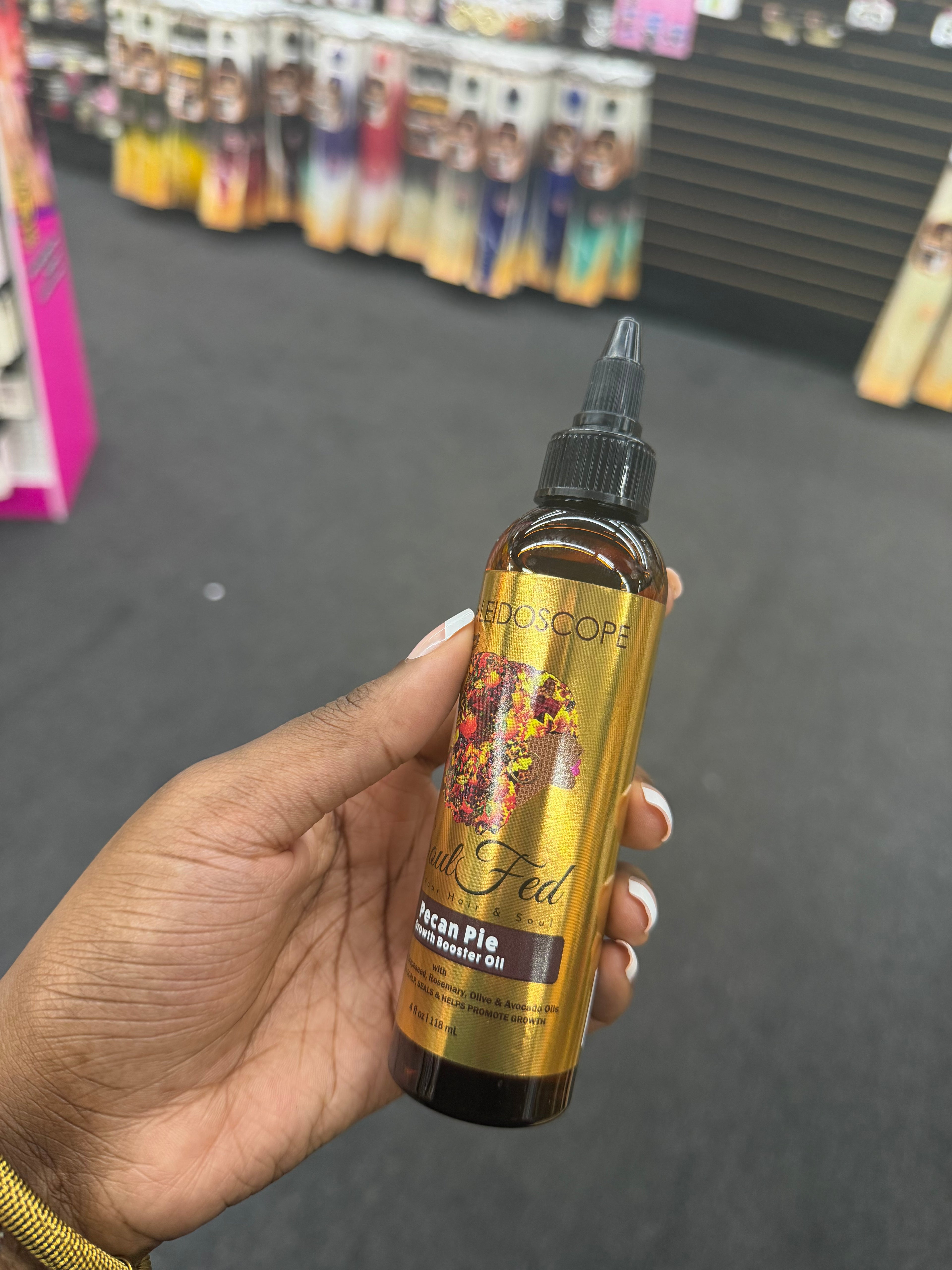 A hand holding a bottle of Alescopa 'A Fan of Pancake' Acian Pie hair serum oil in an amber glass bottle with a gold label and black cap, set against a blurred store background.