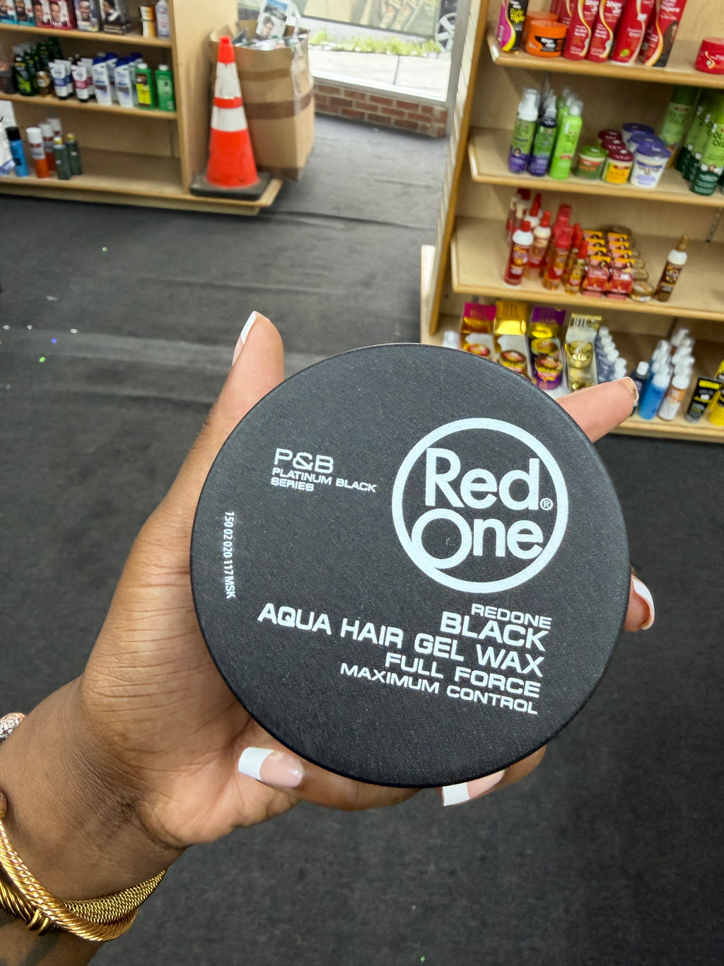 A hand holding a round black container of Red One Aqua Hair Gel Wax in Full Force Maximum Control, with shelves of other products visible in the background of what appears to be a store or salon.