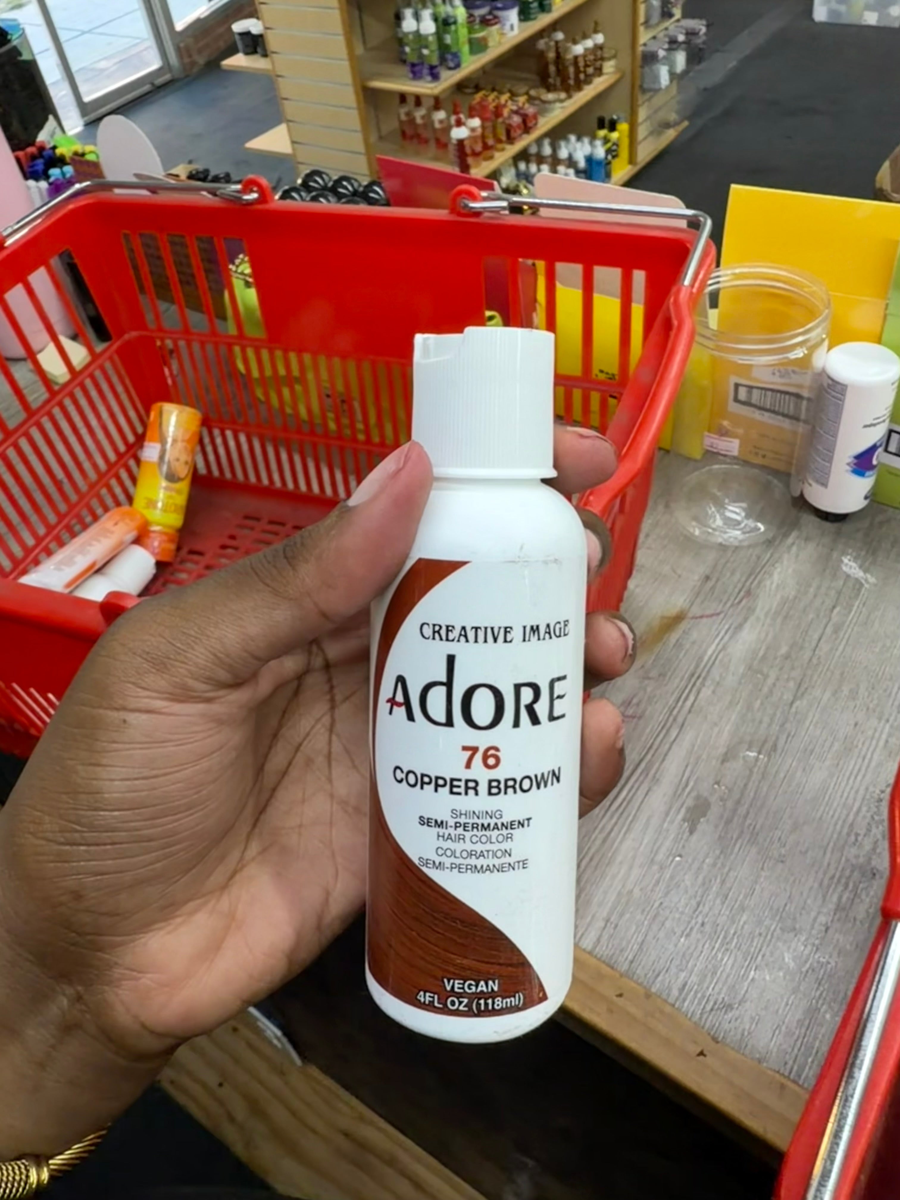 A hand holding a bottle of Creative Image Adore 76 Copper Brown Semi-Permanent Colorant in a retail store setting with a red shopping basket in the background.