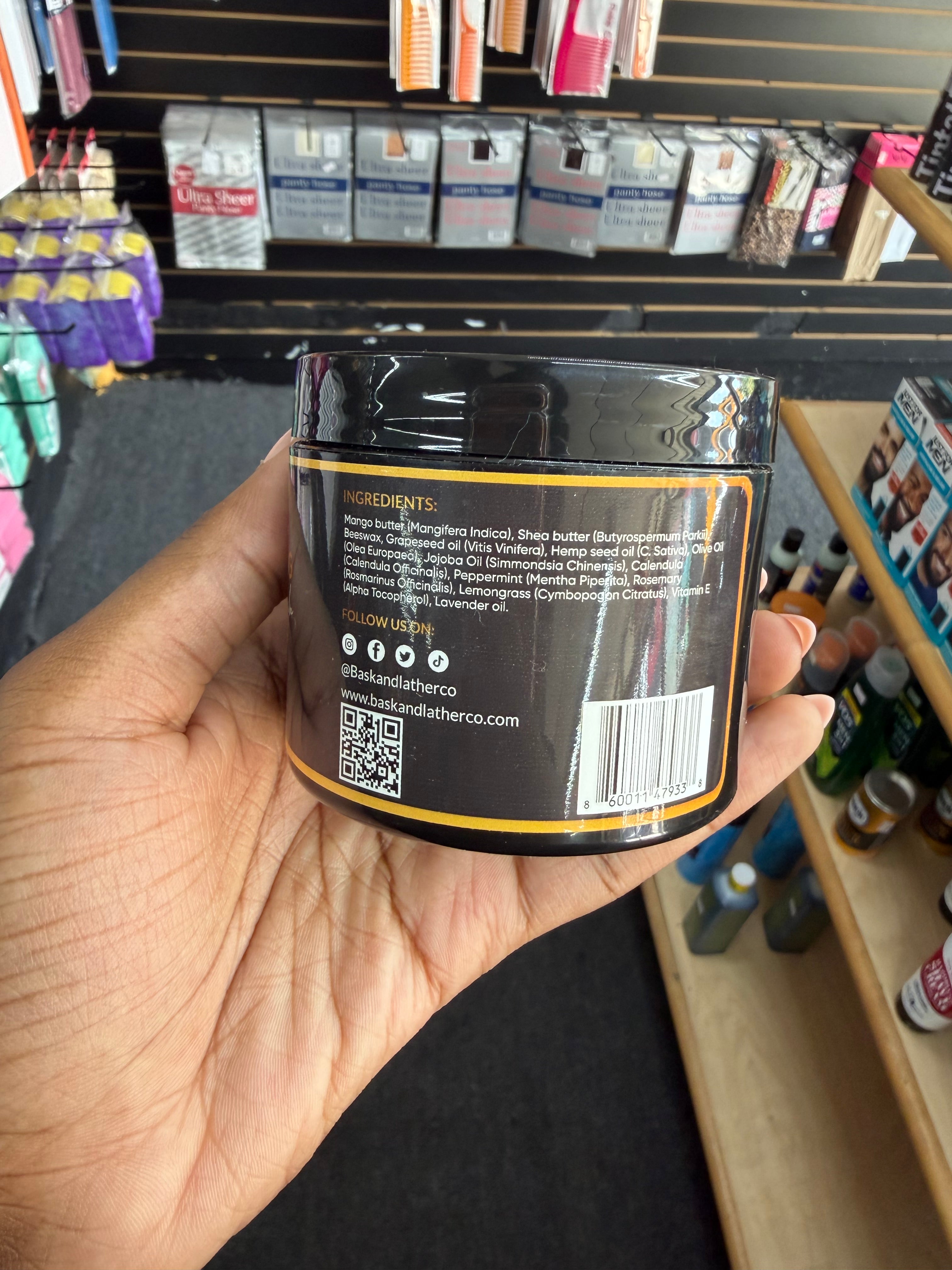 A hand holds a black plastic jar of hair oil in a retail store setting, showing the back label with ingredients and the brand name Showdown.