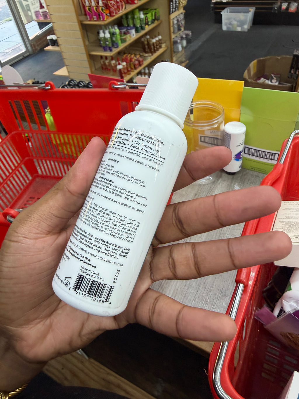 A hand holding a white plastic bottle with a white cap, showing the back label, inside a retail store with red shopping baskets in the background.