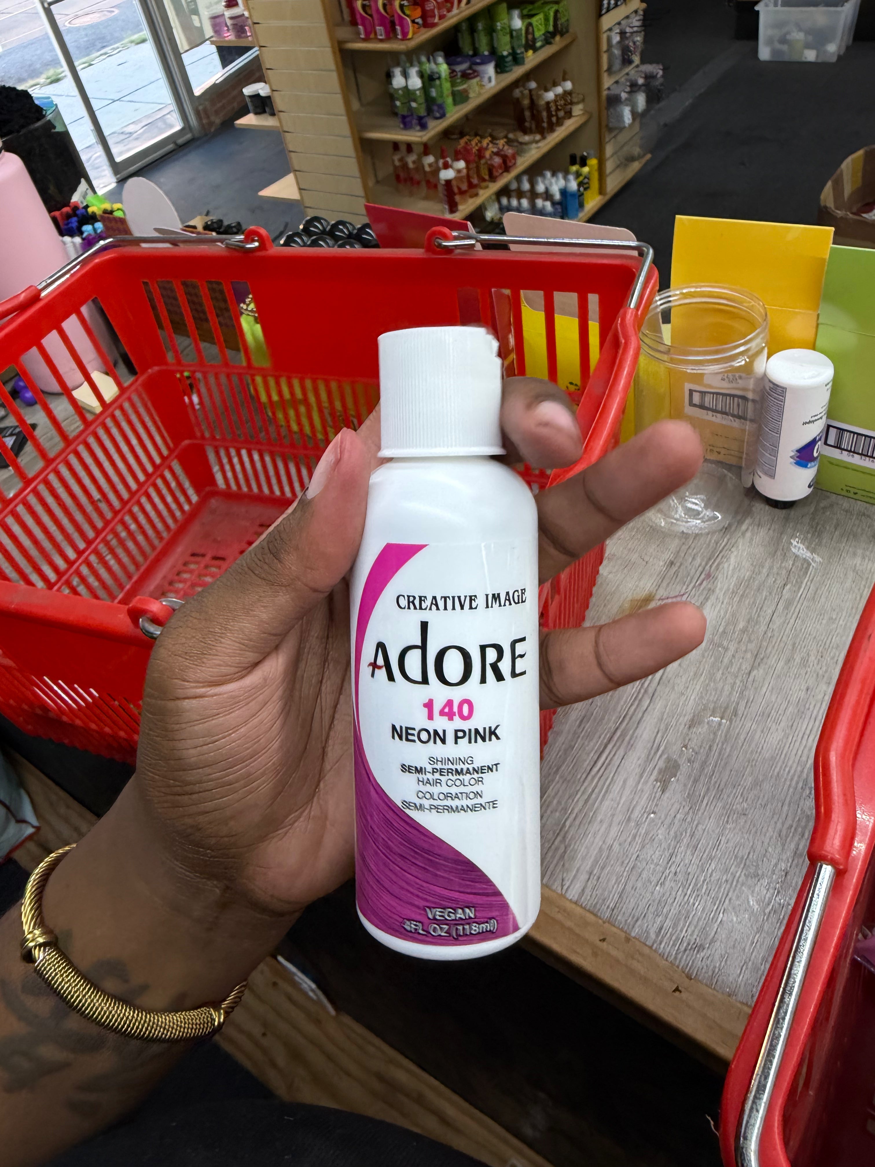 A hand holding a bottle of Creative Image Adore 140 Neon Pink permanent hair color in a retail store setting with a red shopping basket in the background.