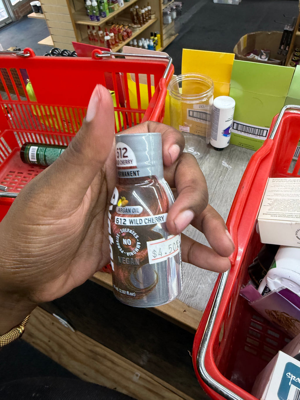 A hand holding a bottle of 512 Wild Cherry organic oil in a grocery store checkout area with red shopping baskets and shelves in the background.