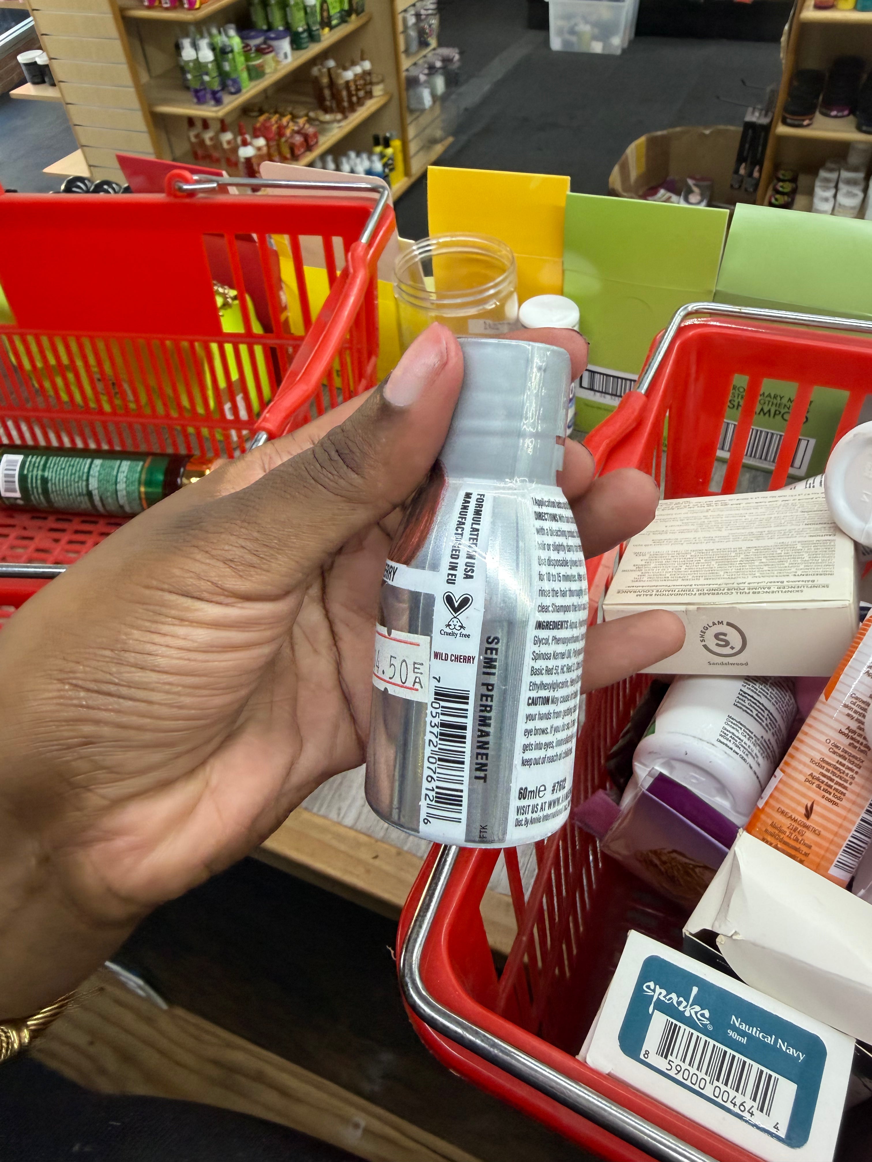 A hand holding a small clear bottle with a white cap and a white product label inside a red shopping basket in a retail store.