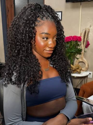 A woman with long, dark, curly hair wearing a gray cardigan and a navy blue crop top, seated indoors with a salon or home interior background featuring pink roses.